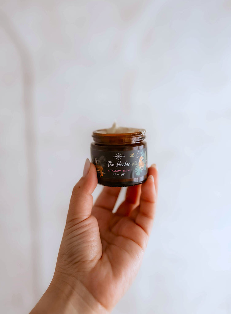 A person holding a jar of "The Healer" from Sun & Moo, showcasing the natural skincare product with a blend of grass-fed tallow, organic baobab oil, and tamanu oil for healing and nourishing the skin.